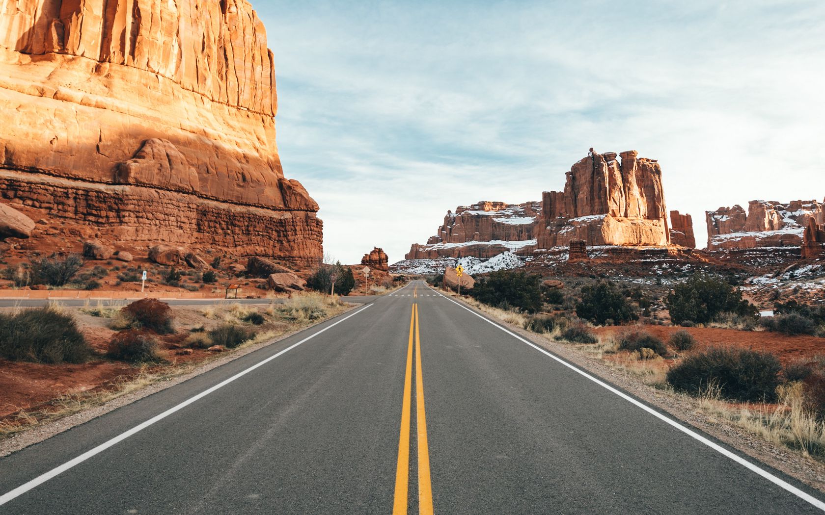 Two-lane asphalt road with double yellow centre lines curving through a desert canyon between tall red sandstone cliff walls